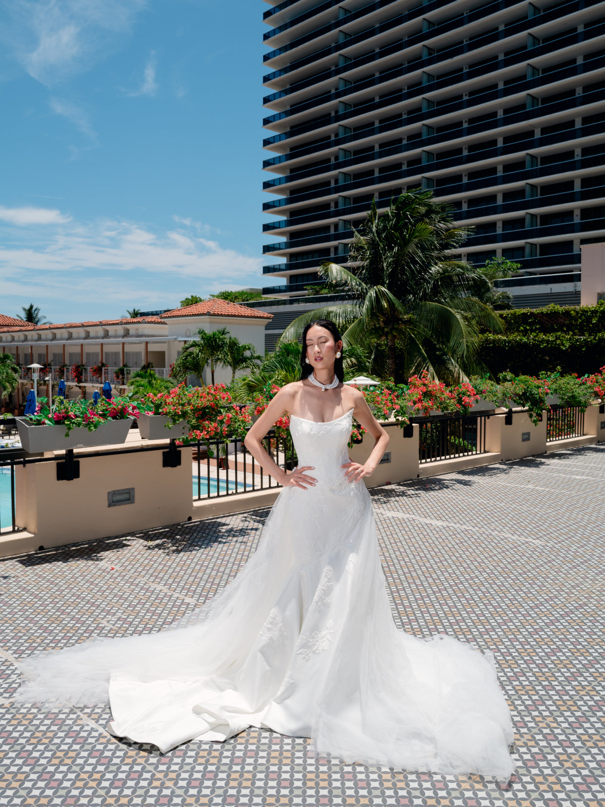 A woman wearing a strapless white wedding gown with a long sheer overskirt and train, standing outdoors on a tiled walkway with a resort pool, palm trees, and a tall hotel building in the background. / Satin mermaid gown with lace appliques and detachable overskirt.