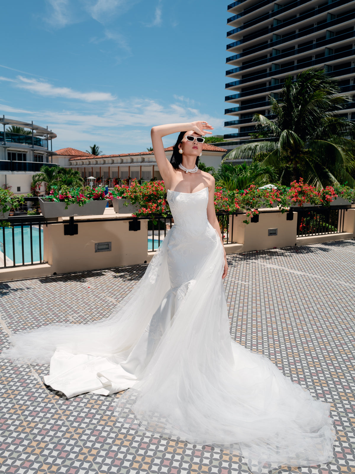 A woman wearing a strapless white wedding gown with a long sheer tulle train stands on a tiled outdoor terrace with palm trees, flowers, and a high-rise building in the background. / Satin mermaid gown with lace appliques and detachable overskirt.