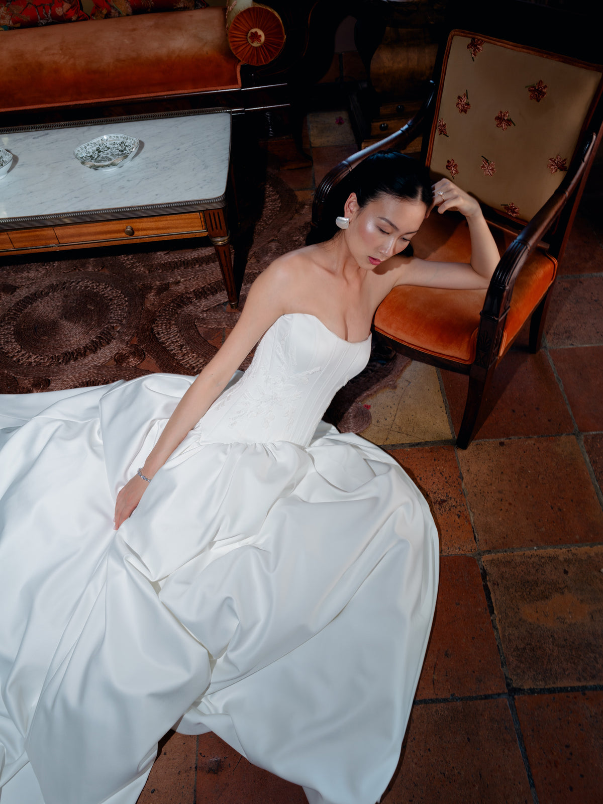 A woman reclining on a terracotta tile floor in a white strapless satin wedding gown, with an orange upholstered chair and a wooden table in the background. / Dramatic drop waist, double face satin ballgown with placed lace and lace up back detail.
