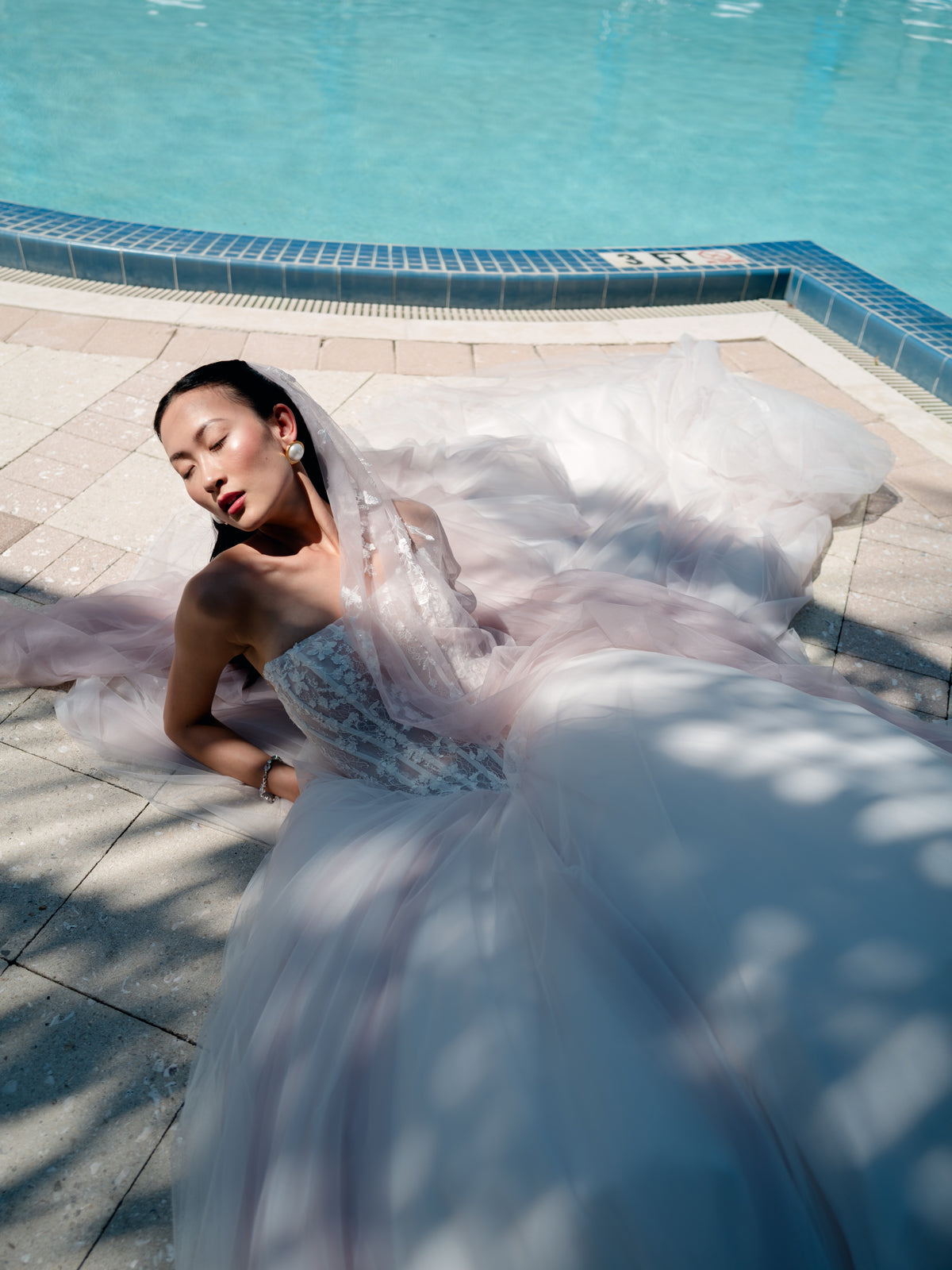 A woman reclines on a tiled poolside deck wearing a voluminous pale blush and white tulle wedding gown with a sheer veil. The bright blue swimming pool and dappled sunlight are visible in the background. / Semi sheer basque waist bodice with lace appliques and dramatic tulle skirt.