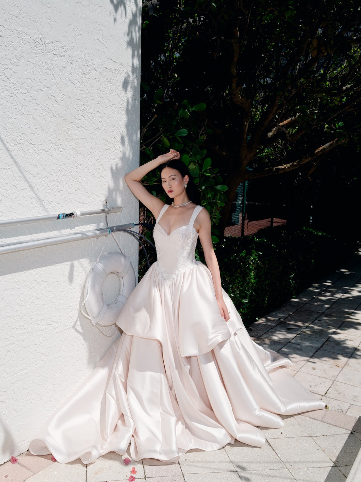 A woman wearing a voluminous ivory satin wedding gown with a sweetheart neckline and layered skirt, posing outdoors against a white stucco wall and green foliage. / Dramatic, sculpted mikado gown with lace placement.
