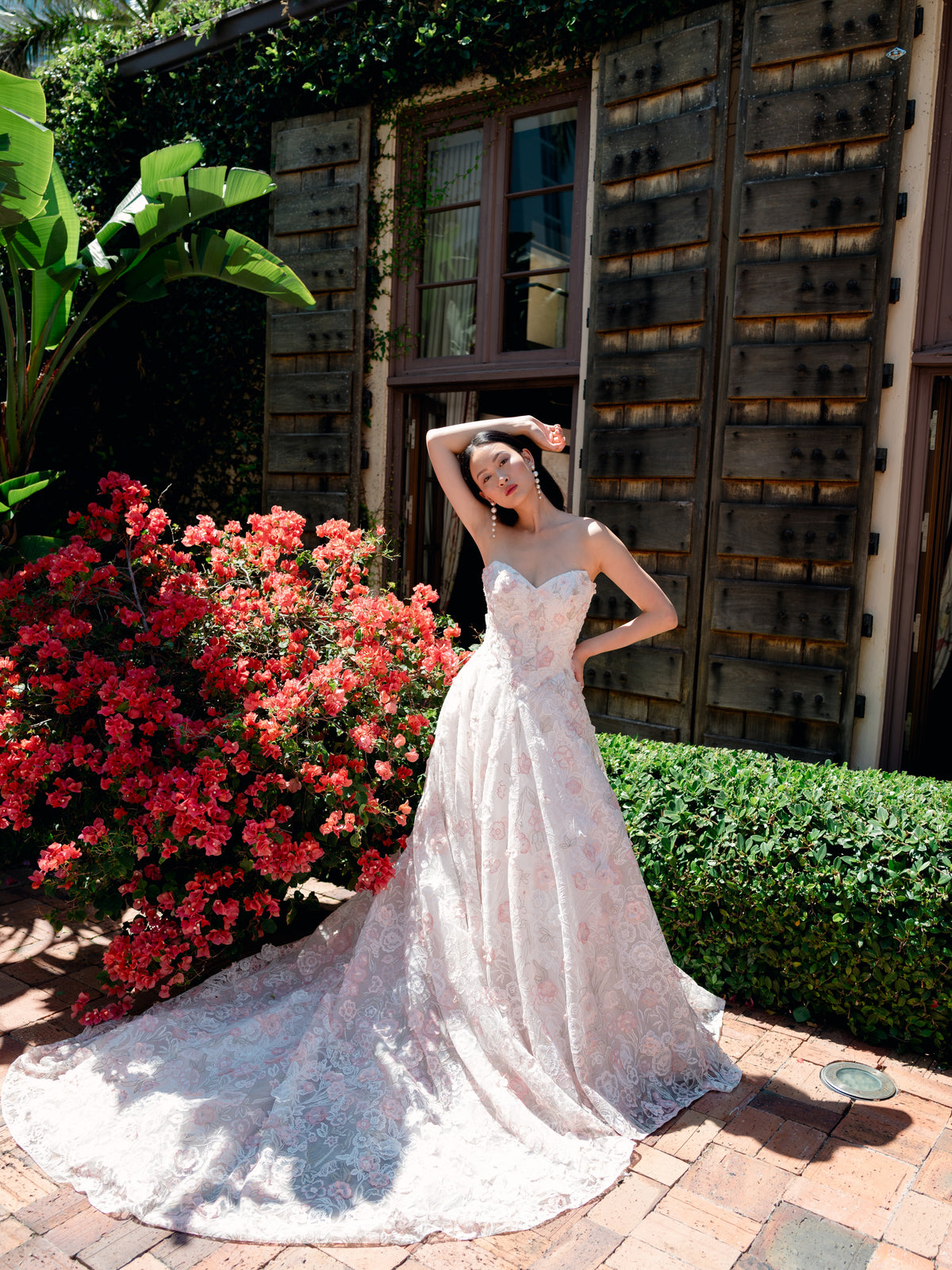 A woman wearing a strapless lace wedding gown with a long train stands outdoors on a brick patio beside a rustic wooden building, red flowering bushes, and green hedges. / All over multicolored lace A-line gown.