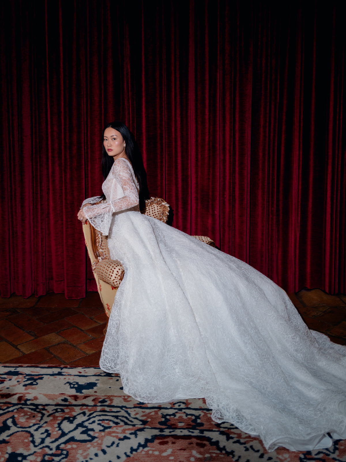 A woman in a voluminous white lace bridal gown with long sleeves sits on a woven chair in front of a dark red velvet curtain. The dress features a dramatic train that spreads across a patterned rug over a wood floor. / All over lace gown with long sleeve, basque waist and A-line skirt with lace up back.