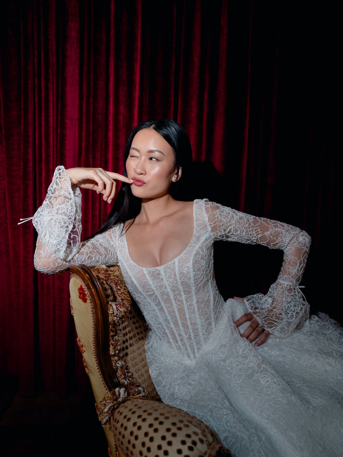 A woman wearing a white lace long-sleeve gown sits on an ornate upholstered chair in front of a dark red velvet curtain. / All over lace gown with long sleeve, basque waist and A-line skirt with lace up back.