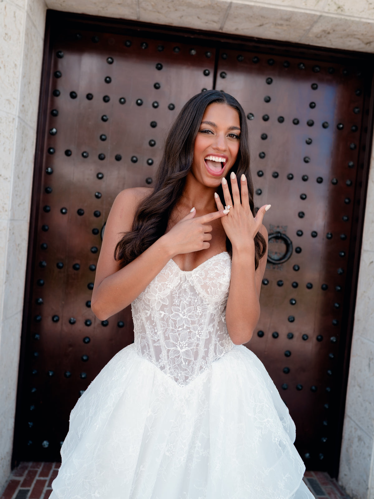 A woman in a white lace wedding gown stands in front of a large dark wooden door, pointing to a ring on her finger with a joyful expression. / All over lace gown with cat eye neckline and soft, dramatic cascading skirt.