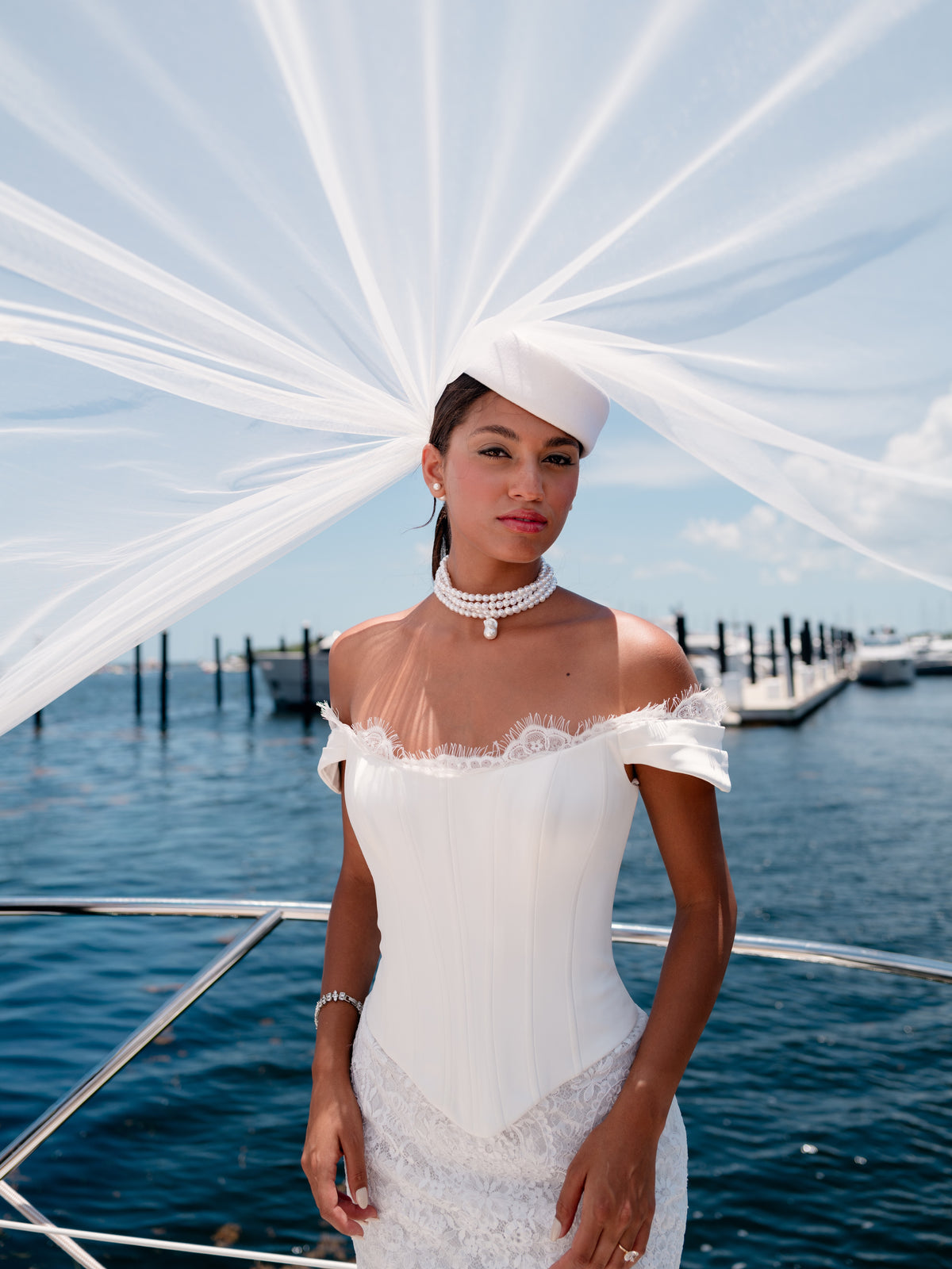 A woman wearing a white off-the-shoulder lace wedding dress and matching white fascinator hat stands on a boat with blue water and sky in the background. / Slim style mock 2 piece with satin bodice and semi-sheer lace skirt. Scooped neckline with off-shoulder straps decorated with delicate lace scallop edge, invisible zipper back closure.