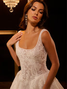 Woman wearing a white lace wedding dress with a chandelier in the background / Ball gown - shimmer tulle with beaded motifs, square neckline with narrow straps finished with fine beaded trim. Buttons/zipper back closure.