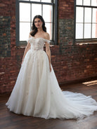 A woman wearing an off-the-shoulder A-line wedding gown with a heavily embellished lace bodice and a long tulle train, standing in a room with exposed brick walls and large windows. / Off the shoulder ballgown with a textured embellishment created with over lace, lace motifs & beaded 3D petals.