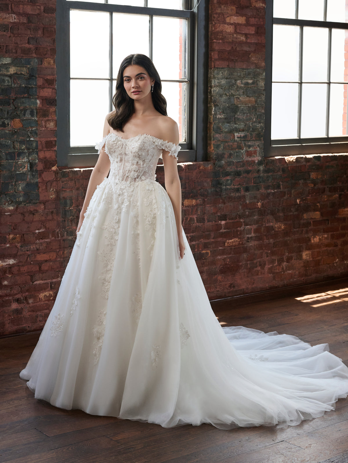 A woman wearing an off-the-shoulder A-line wedding gown with a heavily embellished lace bodice and a long tulle train, standing in a room with exposed brick walls and large windows. / Off the shoulder ballgown with a textured embellishment created with over lace, lace motifs & beaded 3D petals.