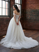 A woman wearing a voluminous off-the-shoulder white wedding gown stands in a brick warehouse interior with large windows, viewed from behind to show the lace-up back and full train. / Off the shoulder ballgown with a textured embellishment created with over lace, lace motifs & beaded 3D petals.