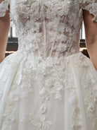 Close-up of a white wedding dress with intricate floral lace applique, beaded detailing, and a full skirt. / Off the shoulder ballgown with a textured embellishment created with over lace, lace motifs & beaded 3D petals.