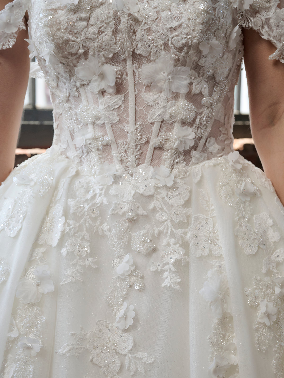 Close-up of a white wedding dress with intricate floral lace applique, beaded detailing, and a full skirt. / Off the shoulder ballgown with a textured embellishment created with over lace, lace motifs & beaded 3D petals.