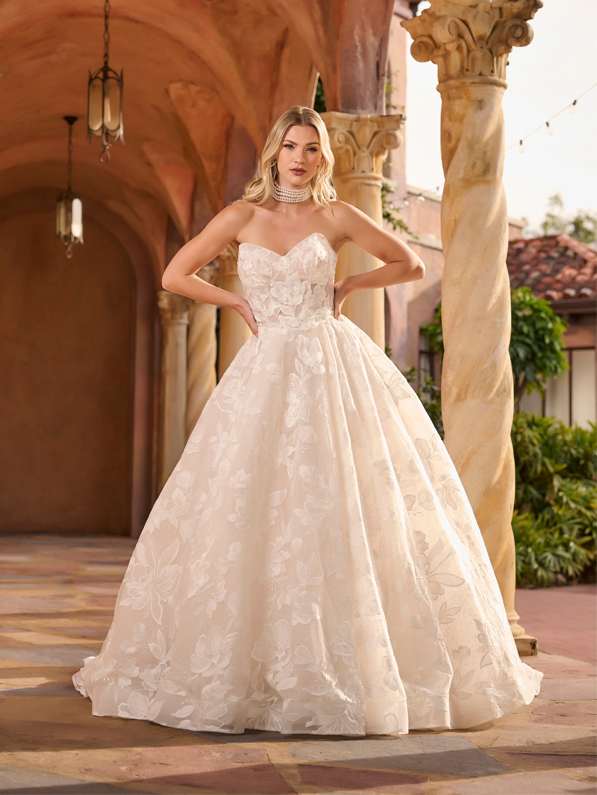 Woman in a white lace wedding dress standing in front of architectural columns. / Full A-line lace gown with sweetheart neckline. Semi-sheer bodice decorated with 3D motifs and beading. Covered buttons and elastic loops along zipper closure at the back.