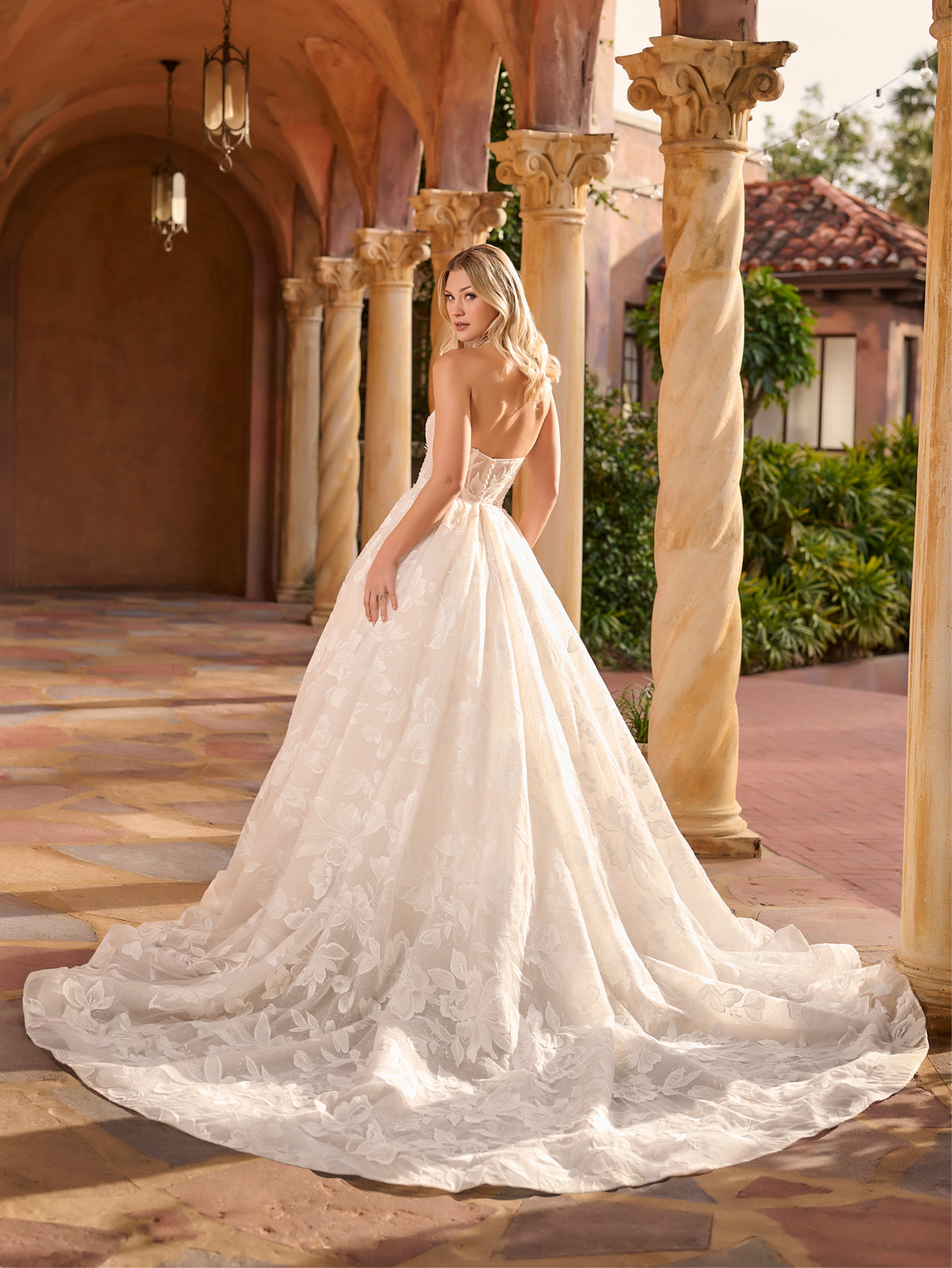 Woman in a white wedding dress standing in an elegant outdoor setting with columns and a building. / Full A-line lace gown with sweetheart neckline. Semi-sheer bodice decorated with 3D motifs and beading. Covered buttons and elastic loops along zipper closure at the back.