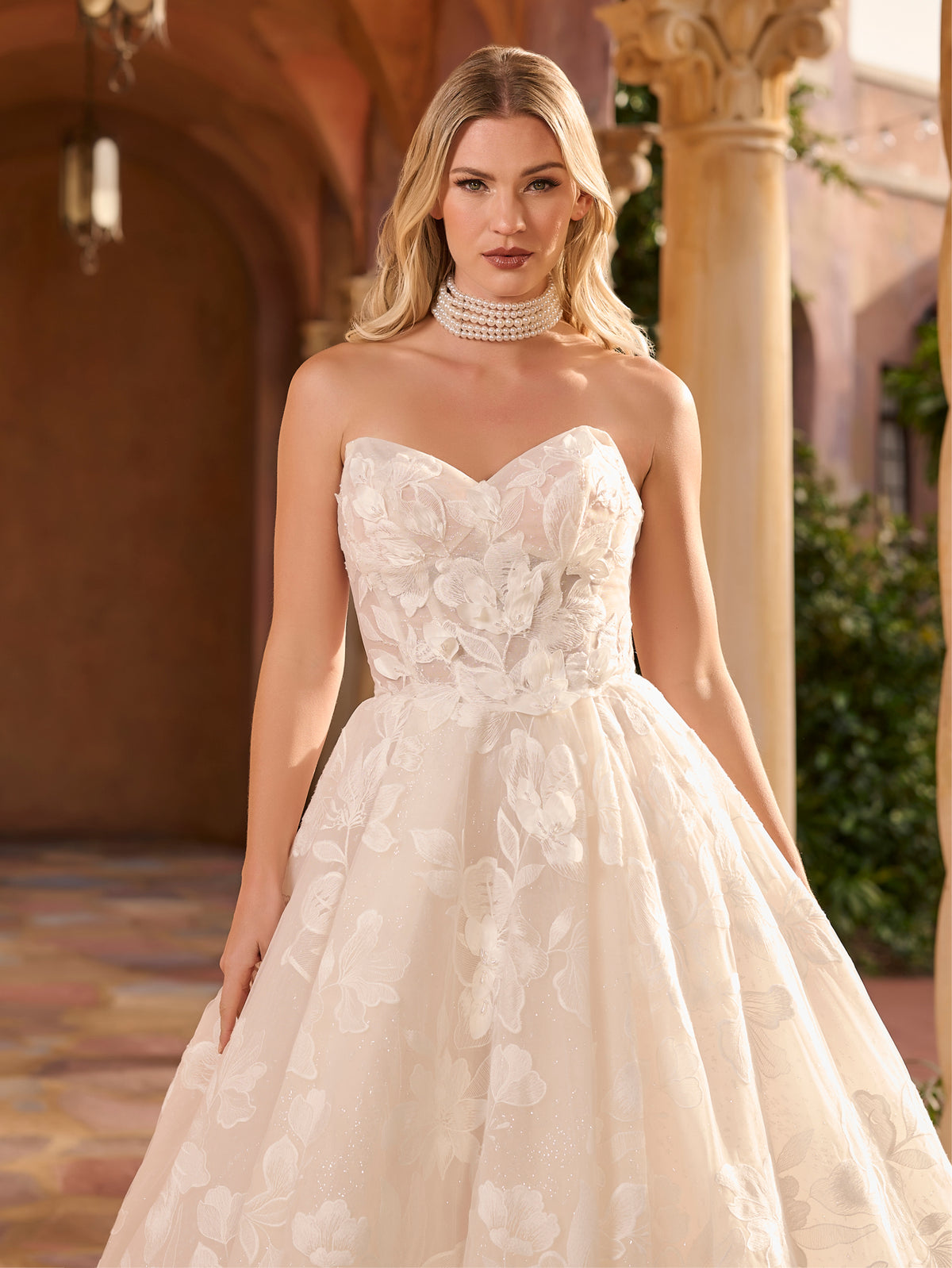 Woman wearing a strapless lace wedding dress in an elegant indoor setting. / Full A-line lace gown with sweetheart neckline. Semi-sheer bodice decorated with 3D motifs and beading. Covered buttons and elastic loops along zipper closure at the back.