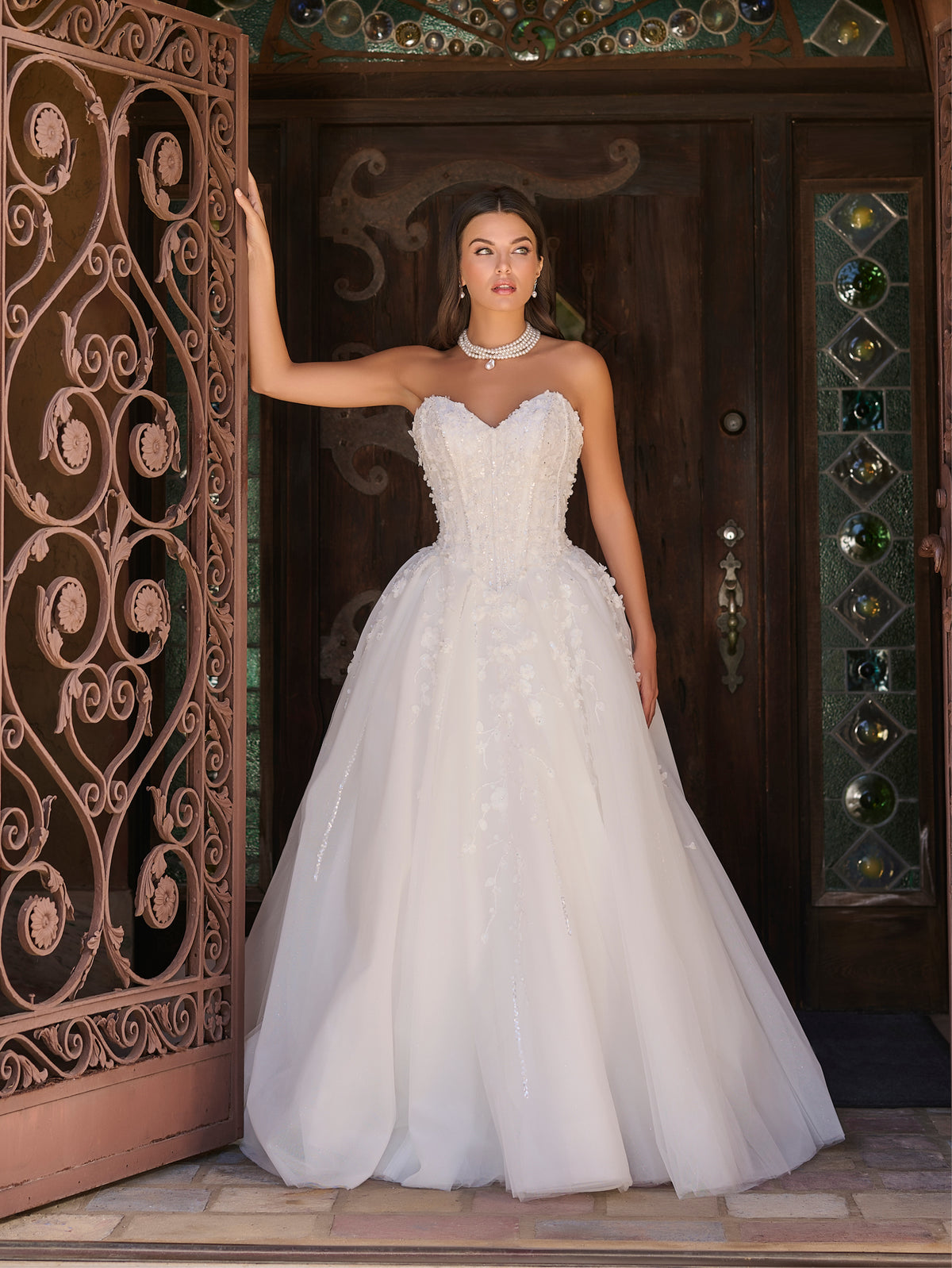 Woman in a white wedding dress standing in front of an ornate wooden door. / Ball gown with sweetheart neckline and exposed boning decorated with hand beading and 3D lace motifs. Lace up back closure.