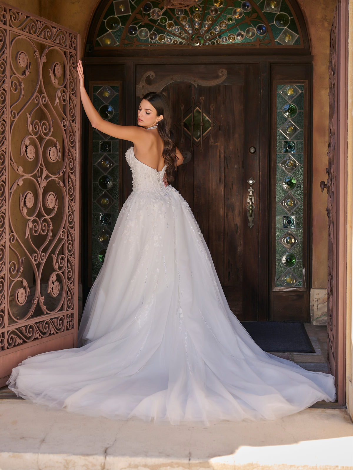 Woman in a white wedding dress standing in front of an ornate wooden door. / Ball gown with sweetheart neckline and exposed boning decorated with hand beading and 3D lace motifs. Lace up back closure.