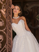 Woman in a white wedding dress standing behind an ornate wooden door. / Ball gown with sweetheart neckline and exposed boning decorated with hand beading and 3D lace motifs. Lace up back closure.