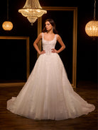 Woman in a white wedding dress standing in a dimly lit room with chandeliers. / Ball gown - shimmer tulle with beaded motifs, square neckline with narrow straps finished with fine beaded trim. Buttons/zipper back closure.