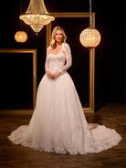 Woman in a white wedding dress standing in a room with chandeliers and framed mirrors. / Ball gown in all over Chantilly lace with detachable lace bolero. Scooped neckline, buttons/zipper back closure.