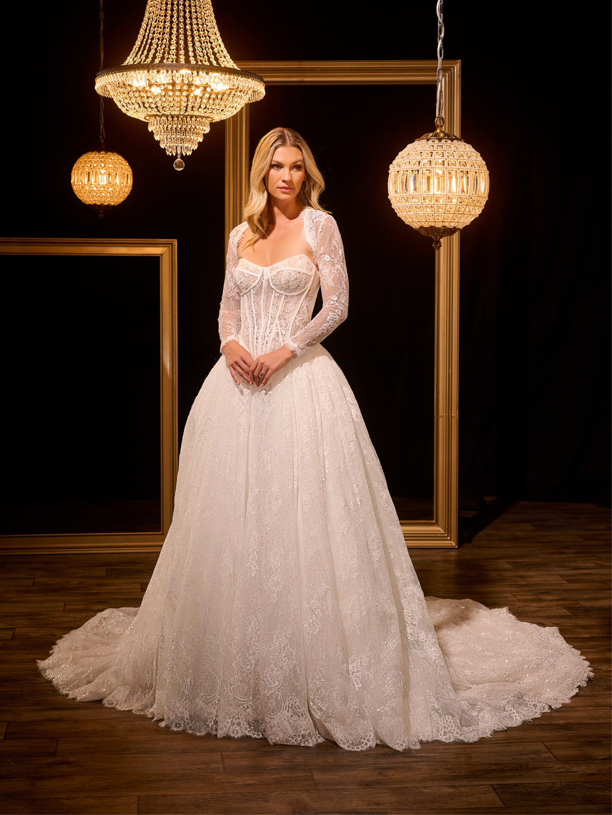 Woman in a white wedding dress standing in a room with chandeliers and framed mirrors. / Ball gown in all over Chantilly lace with detachable lace bolero. Scooped neckline, buttons/zipper back closure.