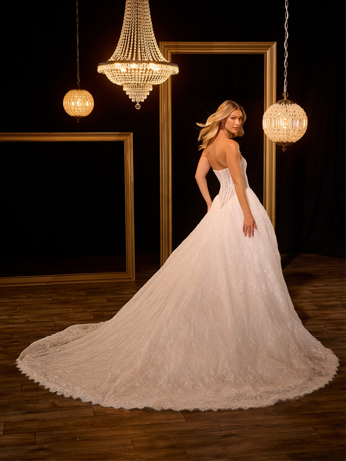 Woman in a white wedding dress standing in a room with chandeliers and framed mirrors. / Ball gown in all over Chantilly lace with detachable lace bolero. Scooped neckline, buttons/zipper back closure.