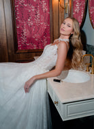 A woman in a white lace and tulle wedding gown leans against a bathroom vanity with a marble sink and gold faucet, set in a room with dark wood paneling and pink floral wallpaper. / Ball gown tulle with lace with scooped neckline and delicate lace straps finished with small bows on front neckline points.