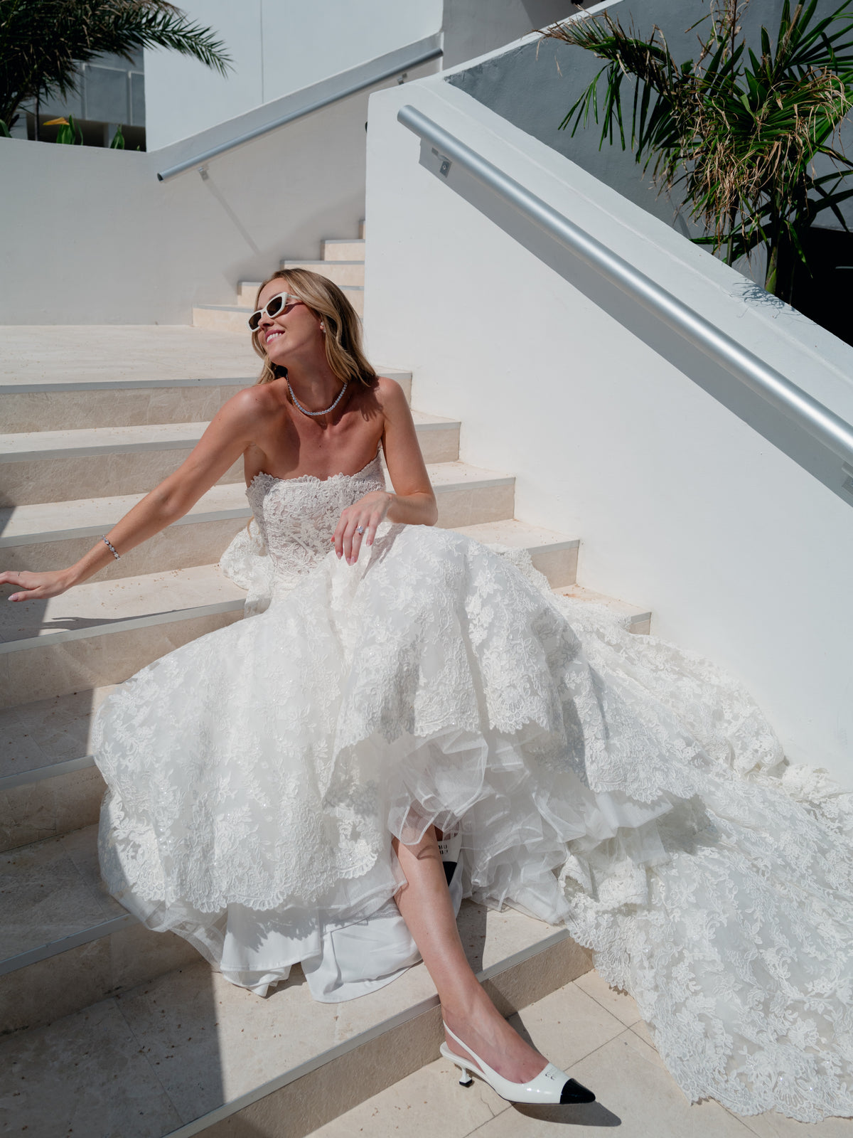 A woman wearing a strapless white lace wedding gown and white pointed-toe heels sits on outdoor stone stairs. The dress has a fitted bodice and a voluminous layered tulle/train, with the model also wearing sunglasses and a necklace. / Mermaid style in all over lace with scooped neckline and lace up back.
