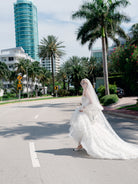 A bride in a voluminous white lace wedding gown and long veil walks across a city street lined with palm trees and modern buildings under a bright sky. / Mermaid style in all over lace with scooped neckline and lace up back.