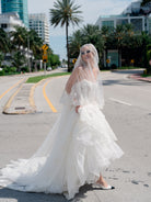 A woman wearing a white strapless wedding gown with a long sheer veil and white pointed flats stands on an urban street with palm trees and buildings in the background. / Mermaid style in all over lace with scooped neckline and lace up back.