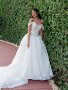 A woman wearing an off-the-shoulder white wedding gown with a lace corset bodice and full tulle skirt, standing outdoors against a green hedge on a reddish paved walkway. / Ball gown with semi-sheer lace bodice and organza skirt. Sweetheart neckline with off-shoulder lace straps. Lace up back closure.