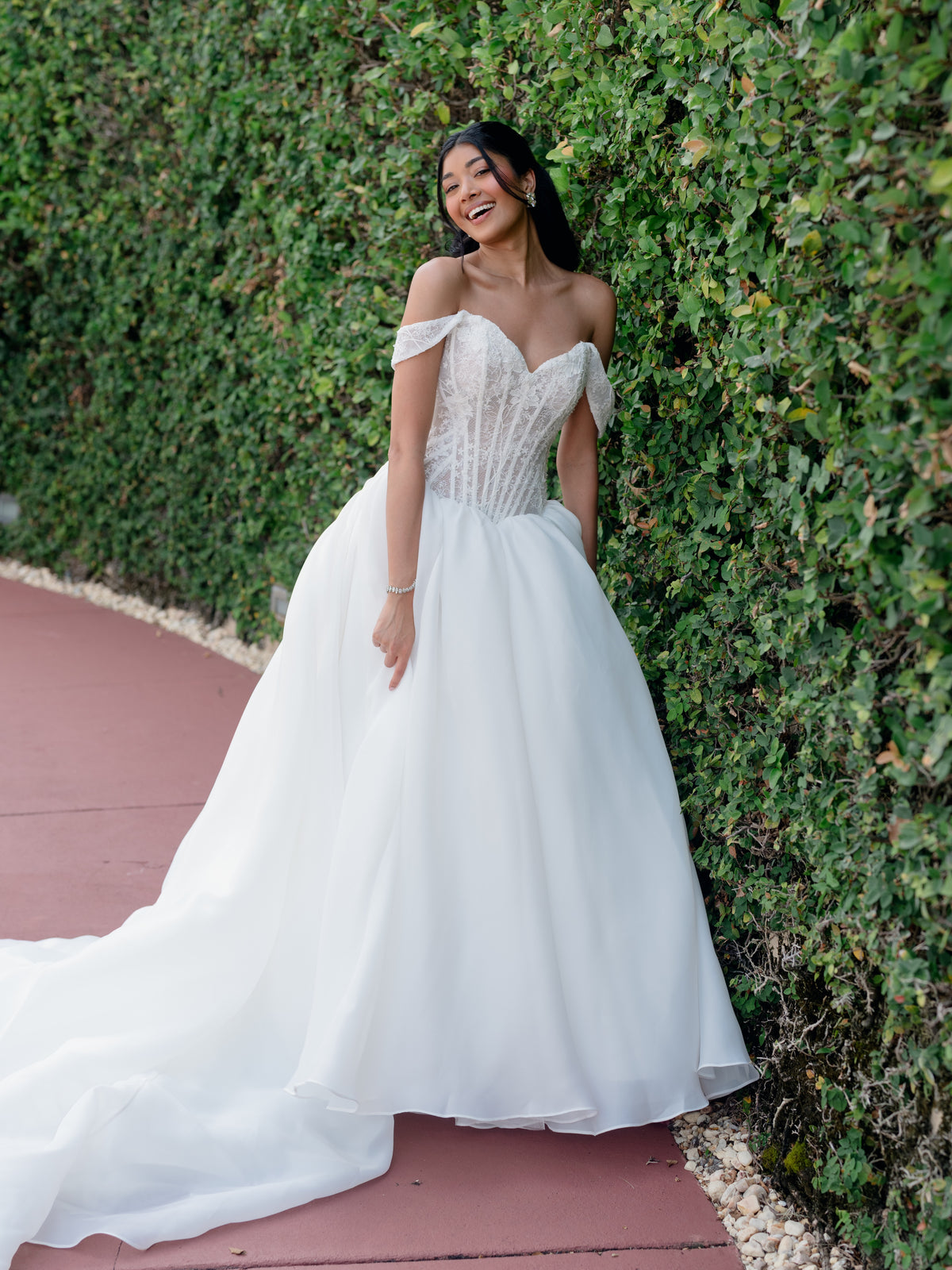 A woman wearing an off-the-shoulder white wedding gown with a lace corset bodice and full tulle skirt, standing outdoors against a green hedge on a reddish paved walkway. / Ball gown with semi-sheer lace bodice and organza skirt. Sweetheart neckline with off-shoulder lace straps. Lace up back closure.