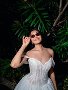 A woman wearing a white off-the-shoulder lace corset gown and dark sunglasses, posing outdoors against a dark background of green foliage. / Ball gown with semi-sheer lace bodice and organza skirt. Sweetheart neckline with off-shoulder lace straps. Lace up back closure.