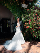 A woman wearing a white wedding dress with a black blazer and a black hat stands outdoors on a brick patio beside a lush green ivy-covered wall with red flowers. / Mermaid lace style with sweetheart neckline and zipper/buttons closure. Detachable tulle skirt with lace motifs applique scattered around the top tulle layer.