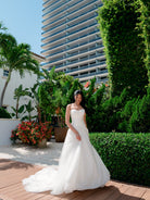 A woman wearing a strapless white wedding gown stands on a wooden deck outdoors, with lush green hedges, potted plants, and a tall modern high-rise building in the background. / Aline tulle gown with drop waist with mikado bodice and tulle skirt, decorated with shimmer lace motifs. Scooped strapless neckline.