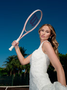 A woman in a white lace tennis dress holds a pink Wilson tennis racket against a clear blue sky with palm trees in the background. / Slim solid lace gown with square neckline and narrow straps. Zipper/buttons back closure.
