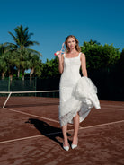 A woman stands on a red clay tennis court wearing a white lace and tulle dress, holding a tennis racket. Palm trees and a blue sky are visible in the background. / Slim solid lace gown with square neckline and narrow straps. Zipper/buttons back closure.