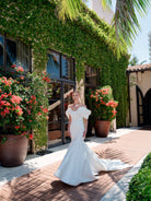 A woman wearing a white off-the-shoulder mermaid-style wedding gown stands on a sunlit walkway in front of a building covered in green ivy, with large potted red flowers and palm leaves visible. / Mermaid style in double face satin with scooped neckline and attached dramatic off-shoulder straps. Bodice decorated with beaded motifs with small 3D flowers. Zipper/buttons back closure.