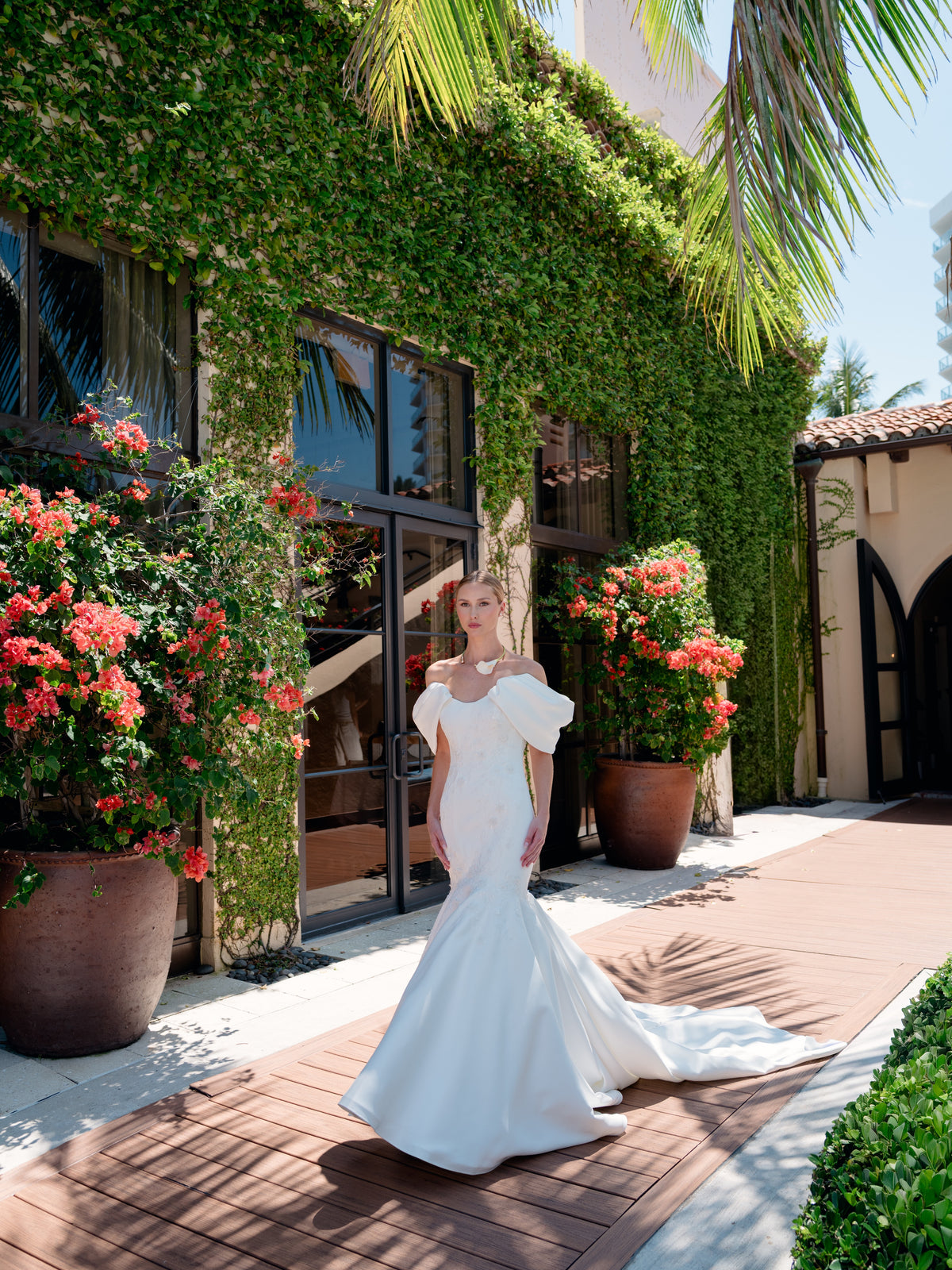 A woman wearing a white off-the-shoulder mermaid-style wedding gown stands on a sunlit walkway in front of a building covered in green ivy, with large potted red flowers and palm leaves visible. / Mermaid style in double face satin with scooped neckline and attached dramatic off-shoulder straps. Bodice decorated with beaded motifs with small 3D flowers. Zipper/buttons back closure.
