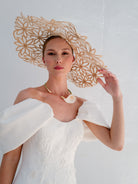 A model wearing a large, ornate beige paper hat with a floral cutout pattern, paired with a white off-the-shoulder dress and a seashell necklace, set against a textured white wall. / Mermaid style in double face satin with scooped neckline and attached dramatic off-shoulder straps. Bodice decorated with beaded motifs with small 3D flowers. Zipper/buttons back closure.