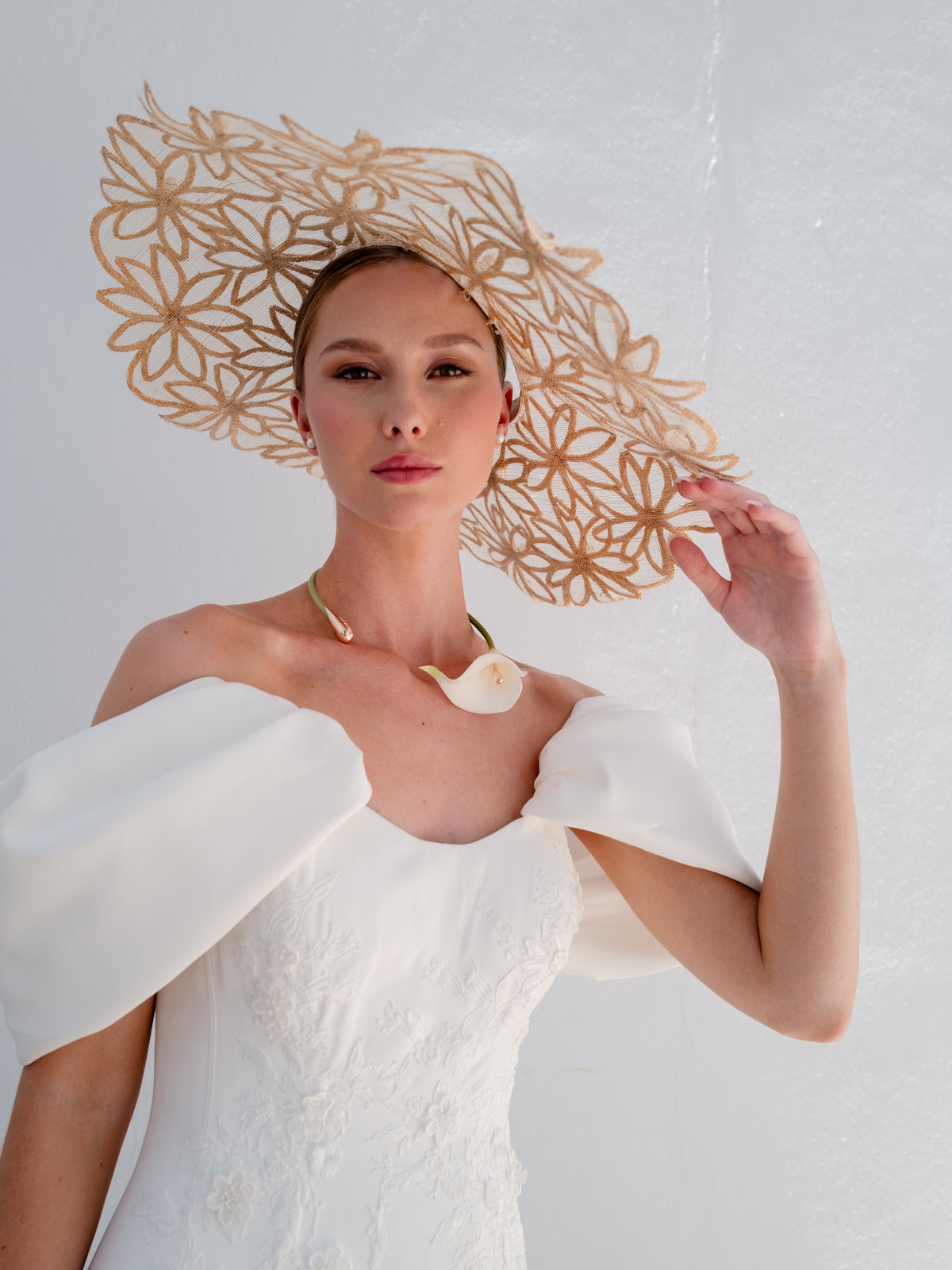 A model wearing a large, ornate beige paper hat with a floral cutout pattern, paired with a white off-the-shoulder dress and a seashell necklace, set against a textured white wall. / Mermaid style in double face satin with scooped neckline and attached dramatic off-shoulder straps. Bodice decorated with beaded motifs with small 3D flowers. Zipper/buttons back closure.