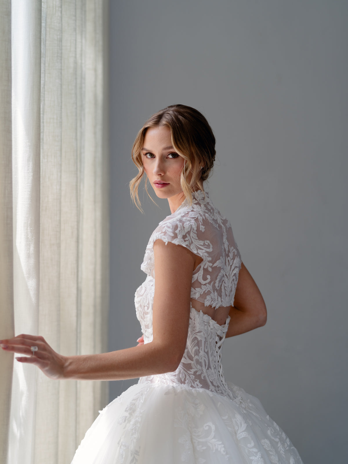 A woman wearing a white lace wedding dress stands in profile near a window with sheer curtains, looking back over her shoulder. The dress has a fitted lace bodice, sheer cap sleeves, and a full tulle skirt. / Ball gown with semi sheer bodice with scooped neckline decorated with beaded lace motifs scattered down onto the skirt. Lace up back closure. Matching lace bolero with cup sleeves.