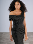 Woman wearing a black off-shoulder dress with floral embellishments against a gray background / Slim gown with side draping and asymmetrical shoulder and off-shoulder strap on one side. The dress is decorated with beautiful, beaded motifs on the right front side and features a side slit.