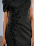 A close-up of a black draped evening gown with a one-shoulder design and a vertical line of black beaded floral embellishments on the side, worn by a model. / Slim gown with side draping and asymmetrical shoulder and off-shoulder strap on one side. The dress is decorated with beautiful, beaded motifs on the right front side and features a side slit.