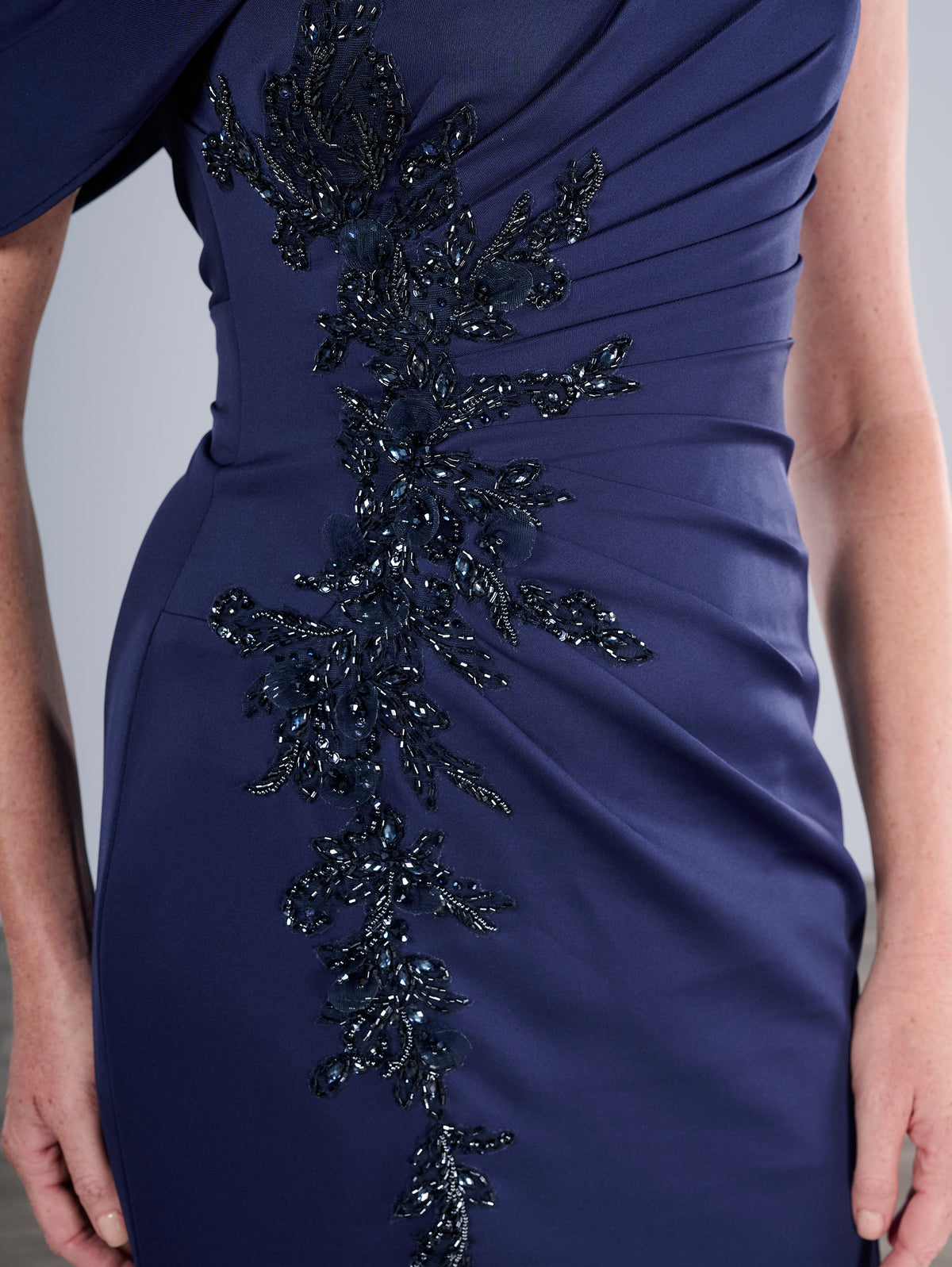 Close-up of a navy blue evening gown with a vertical column of dark beaded embellishment running down the front, shown on a model. / Slim gown with side draping and asymmetrical shoulder and off-shoulder strap on one side. The dress is decorated with beautiful, beaded motifs on the right front side and features a side slit.