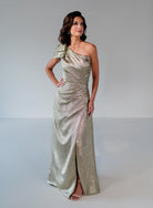 A woman modeling a floor-length, one-shoulder metallic champagne gown with a shimmering textured fabric, set against a plain light gray studio background. / This one shoulder metallic brocade gown features a pleated bodice with a wrap skirt.