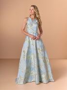 A woman modeling a sleeveless halter-neck A-line gown in light blue with a gold floral pattern, set against a solid warm beige studio background. / This stunning gown features a softly muted brocade fabric with a halter neckline and a belted waist.