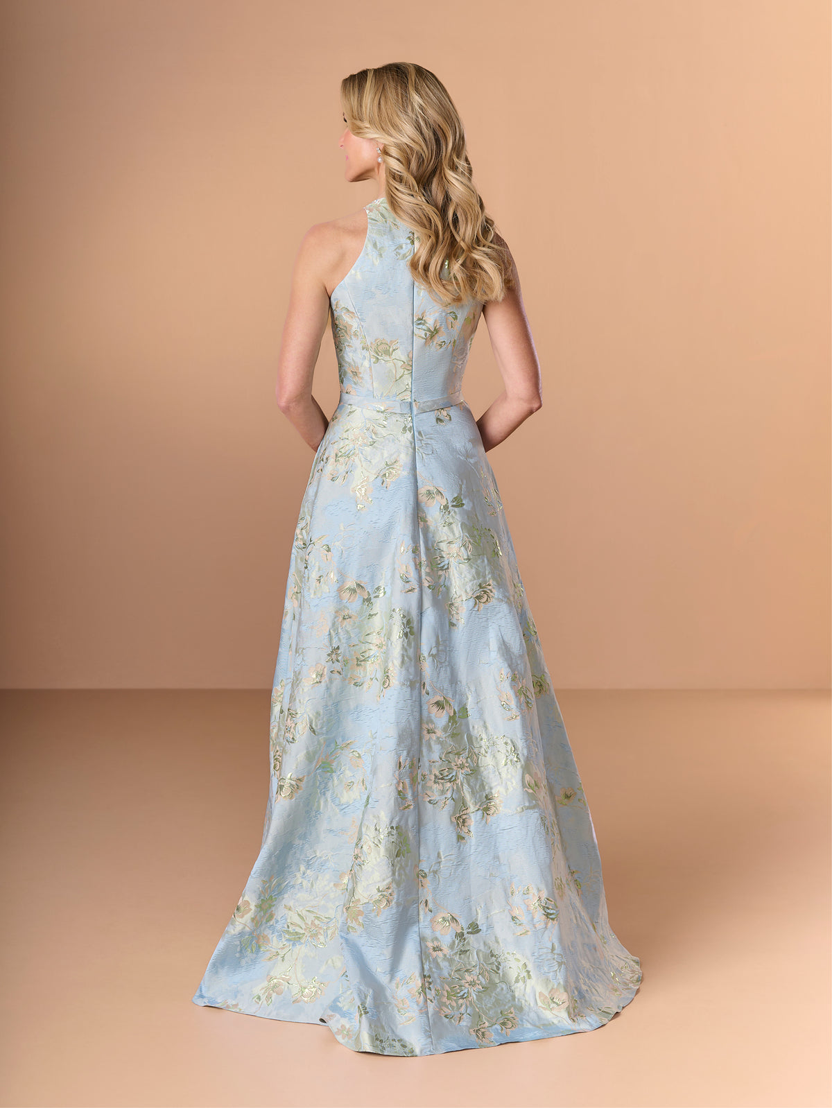 A woman modeling a light blue halter-neck floor-length gown with a gold floral pattern, shown from the back against a solid peach studio backdrop. / This stunning gown features a softly muted brocade fabric with a halter neckline and a belted waist.
