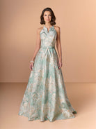 A woman modeling a sleeveless halter-neck floor-length gown with a light aqua and beige floral jacquard pattern, set against a solid warm beige studio background. / This stunning gown features a softly muted brocade fabric with a halter neckline and a belted waist.