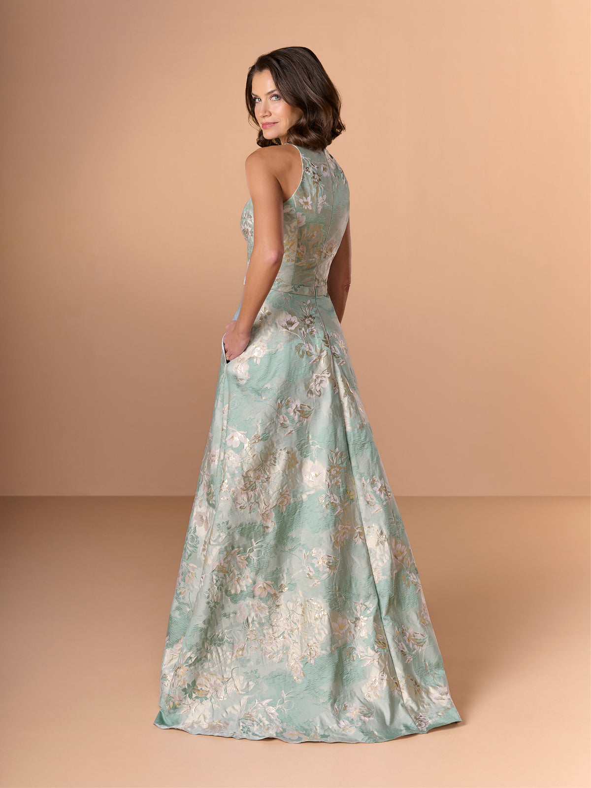 A woman modeling a floor-length formal gown with a mint green and beige floral jacquard pattern, shown from a back-three-quarter angle against a solid warm beige studio backdrop. / This stunning gown features a softly muted brocade fabric with a halter neckline and a belted waist.